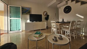 A modern living room with a flat-screen TV, a dining area with a wooden table and chairs, and a small round table with cups and books. - Cozy Home With Terrace and Access to Swimming Pool! (Mogan)