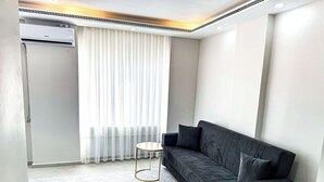 Una sala de estar moderna con un sofá gris oscuro, una pequeña mesa redonda y un amplio ventanal con cortinas traslúcidas.