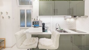 A modern kitchen with white cabinetry, a white countertop, and a built-in stove. There are two white chairs with fluffy cushions, a sink, and a window with frosted glass. - Margaret Studio by the Danube near Margaret Island (Budapest)