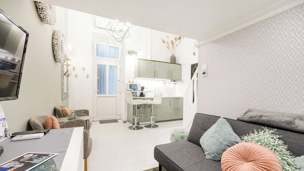 A modern living room with a grey sofa, a coffee table, and a TV. A kitchen is visible in the background with white cabinets and a window. - Margaret Studio by the Danube near Margaret Island (Budapest)