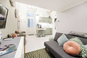 Una sala moderna con un sofá gris, una mesa de centro y un televisor. Se ve una cocina al fondo con armarios blancos y una ventana.