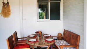 A covered outdoor dining area with a wooden table and chairs, a ceiling fan, and a window with a view of greenery. - Following the Horizon of the Balisiers (Sainte-Anne)