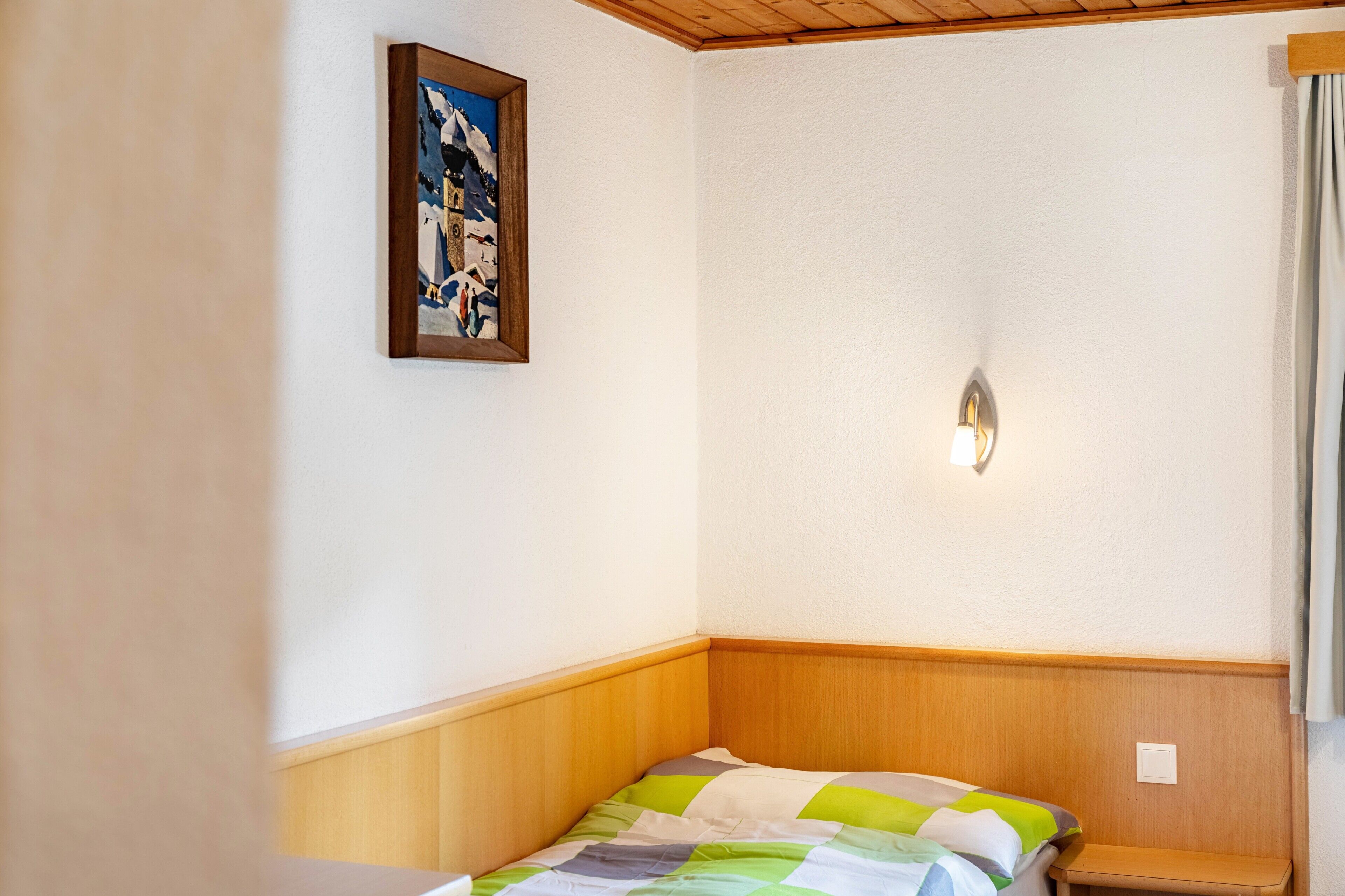 A corner bedroom with a wooden headboard, a bed with a green and white striped bedspread, a framed picture on the wall, and a light fixture.