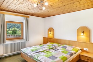 Een slaapkamer met een houten plafond, een bed met een geruit beddegoed, een nachtkastje, een wandlamp en een raam met gordijnen.