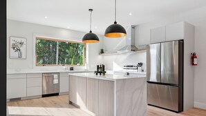 A modern kitchen with a large island, stainless steel appliances, pendant lighting, and a view of the outdoors. - Modern Muskoka River Retreat (Bracebridge)