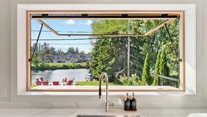 A kitchen with a large window overlooking a lake, outdoor seating, and a stainless steel sink. - Modern Muskoka River Retreat (Bracebridge)