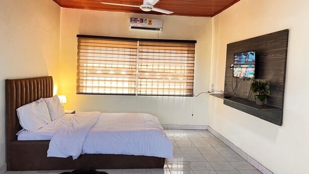 Un dormitorio con cama, ventilador de techo, equipo de aire acondicionado, ventana con persianas y televisor montado.