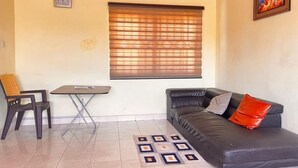 Una sala de estar con sofá, una mesita, una silla, una ventana con persianas y un reloj en la pared.
