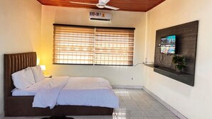 Un dormitorio con cama, ventilador de techo, ventana con persianas, televisor montado en la pared y una planta pequeña.