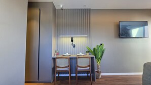 A modern dining area with a grey wall, a flat-screen TV mounted above a table with two wicker chairs, a potted plant, and a wall-mounted phone.