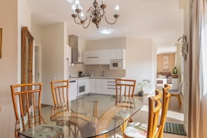 A dining area with a glass table surrounded by wooden chairs, a kitchen in the background, and a chandelier.