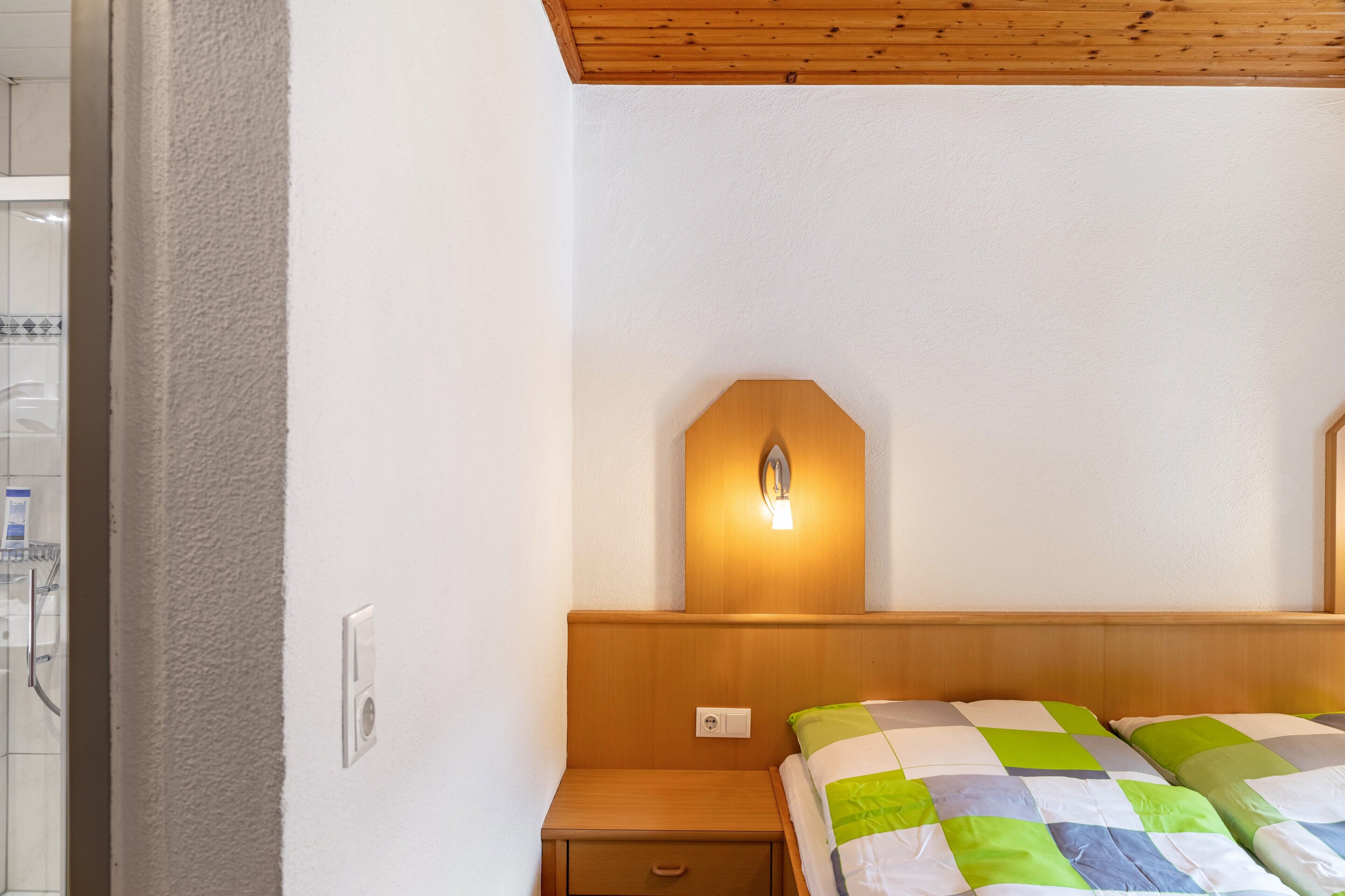 Quarto com cabeceira de madeira, luminária de cabeceira, colcha acolchoada e mesa de cabeceira com gaveta.