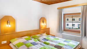 Un dormitori amb sostre de fusta, un llit amb colcha escacada, dues làmpades fixades a la paret i una finestra amb cortines.