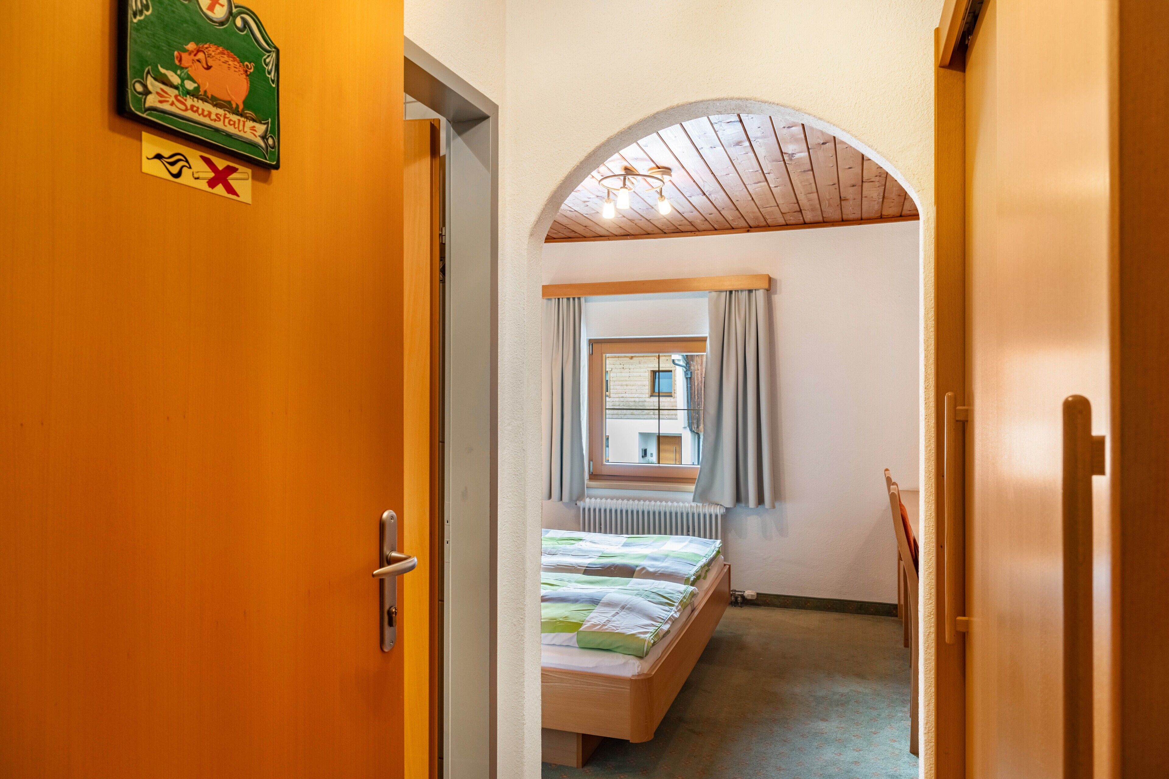 Passadís estret que condueix a una habitació amb un llit, una finestra amb cortines i un sostre de fusta.