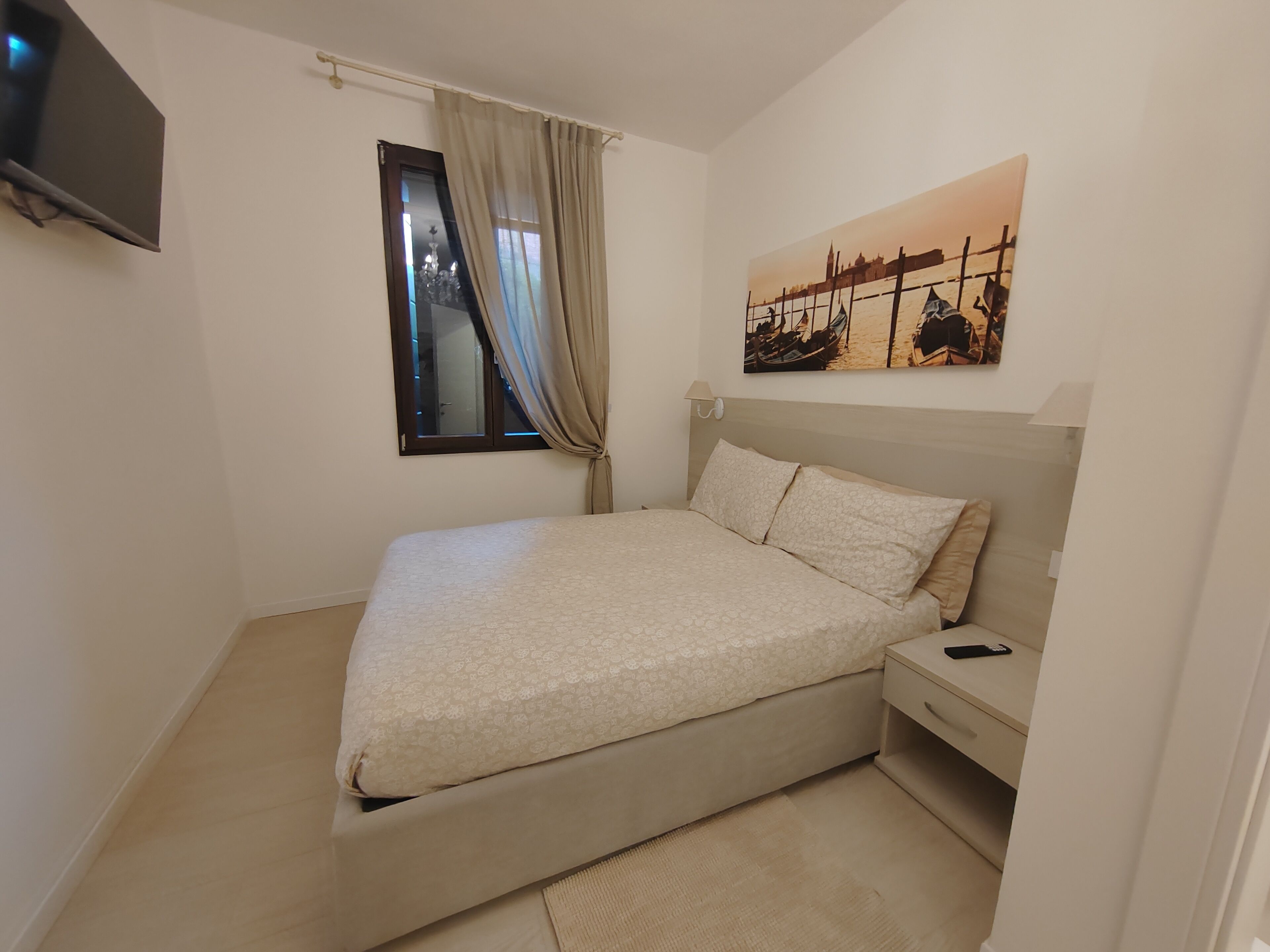 A bedroom with a bed, a nightstand, a TV, a window with curtains, and a painting on the wall.