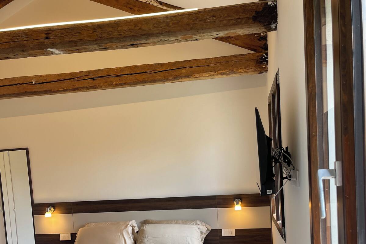 A bedroom with a bed, wooden beams, a wall-mounted TV, and a window.