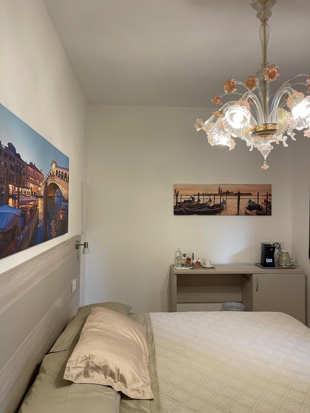 A hotel room with a bed, a desk with a coffee maker, and two paintings on the wall.