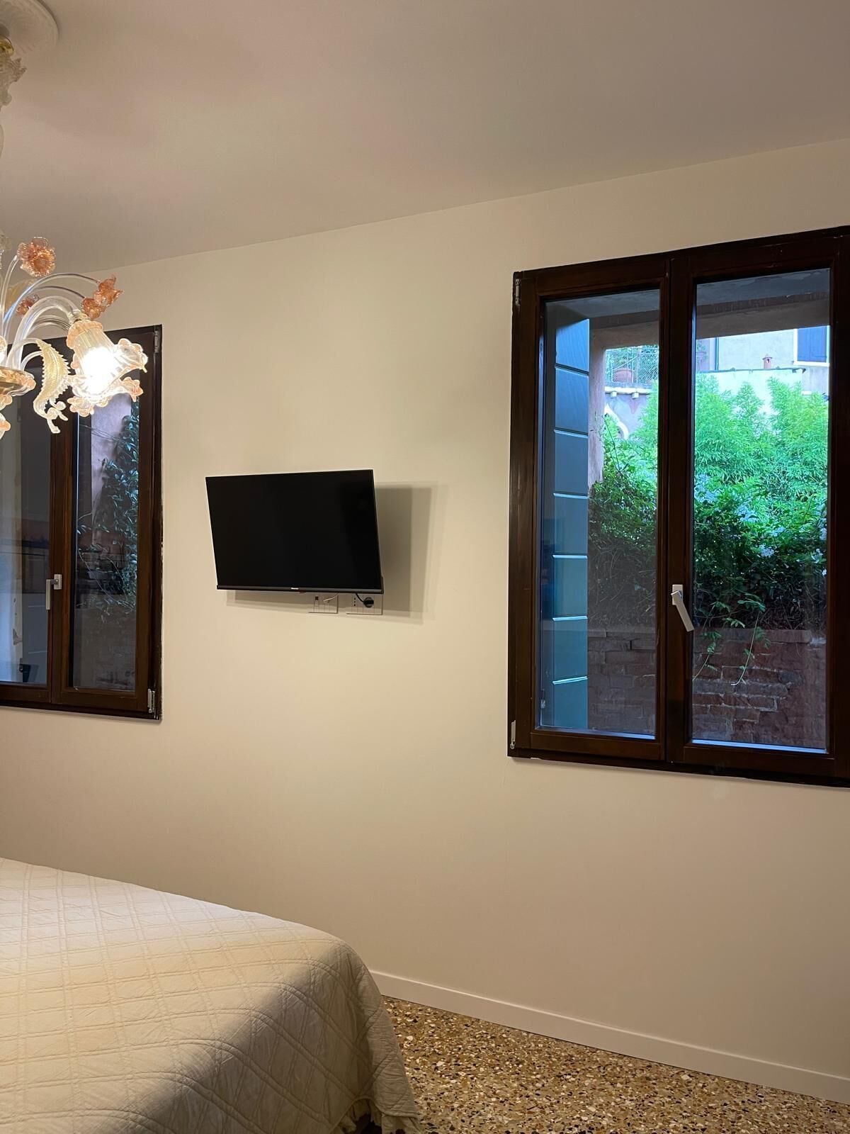 A bedroom with a bed, a TV mounted on the wall, and a window with a view of greenery outside.