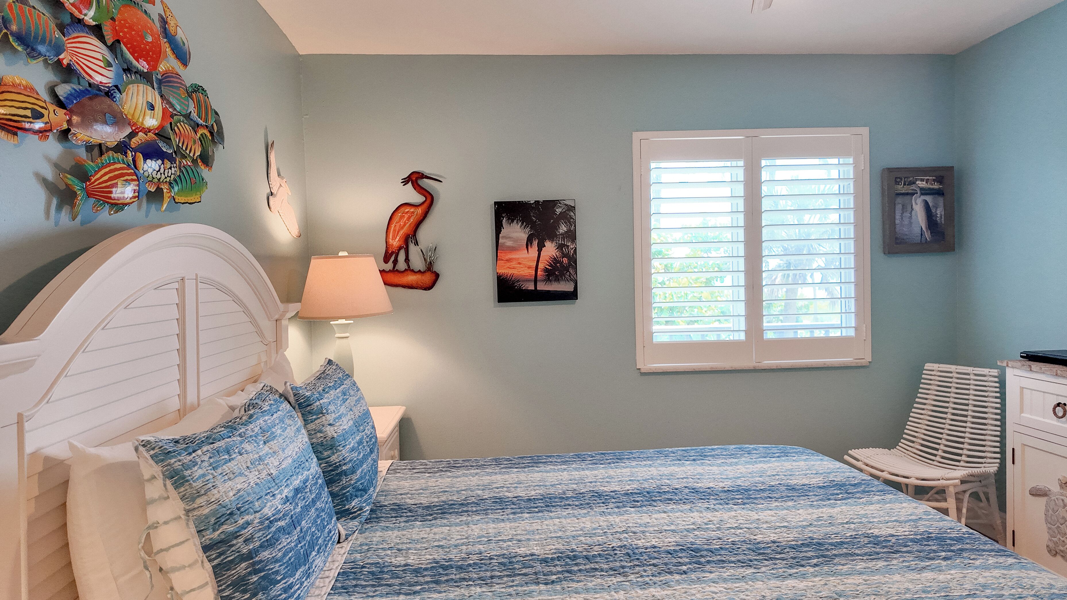 A bedroom with a bed, a nightstand, a lamp, a window with blinds, and wall art including a bird and a sunset scene.