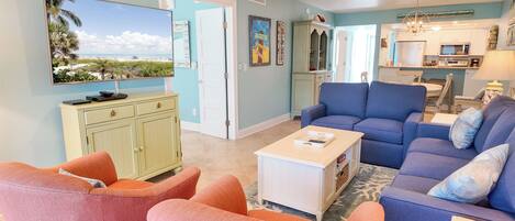 A living room with a TV, a blue sofa, orange chairs, and a kitchen in the background.