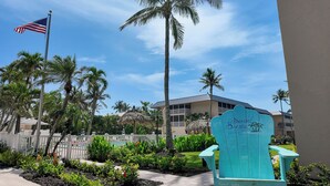 Egy tengerparti szálloda, amelyen egy „Sanibel Siesta On the Beach” feliratú tábla látható, és amelyet pálmafák és egy fehér, deszkából készült kerítés vesz körül.