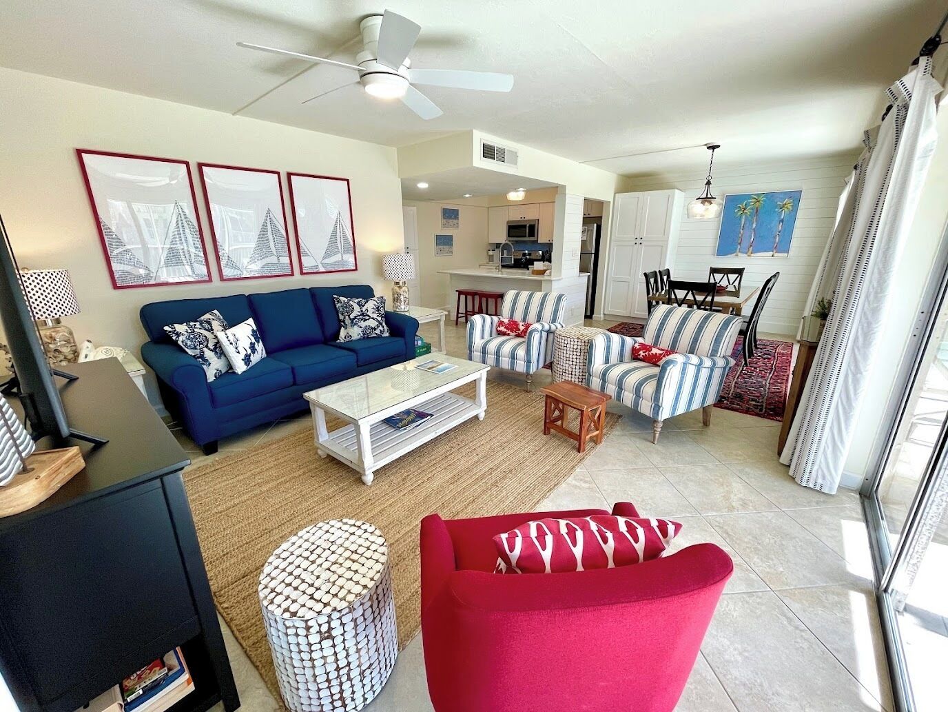 A living room with a blue sofa, a red armchair, a coffee table, and a patterned ottoman. There is a ceiling fan, a dining area with striped chairs, and a kitchen in the background.