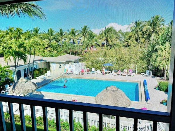 A pool area with lounge chairs and umbrellas, surrounded by palm trees and a building with a sloped roof.