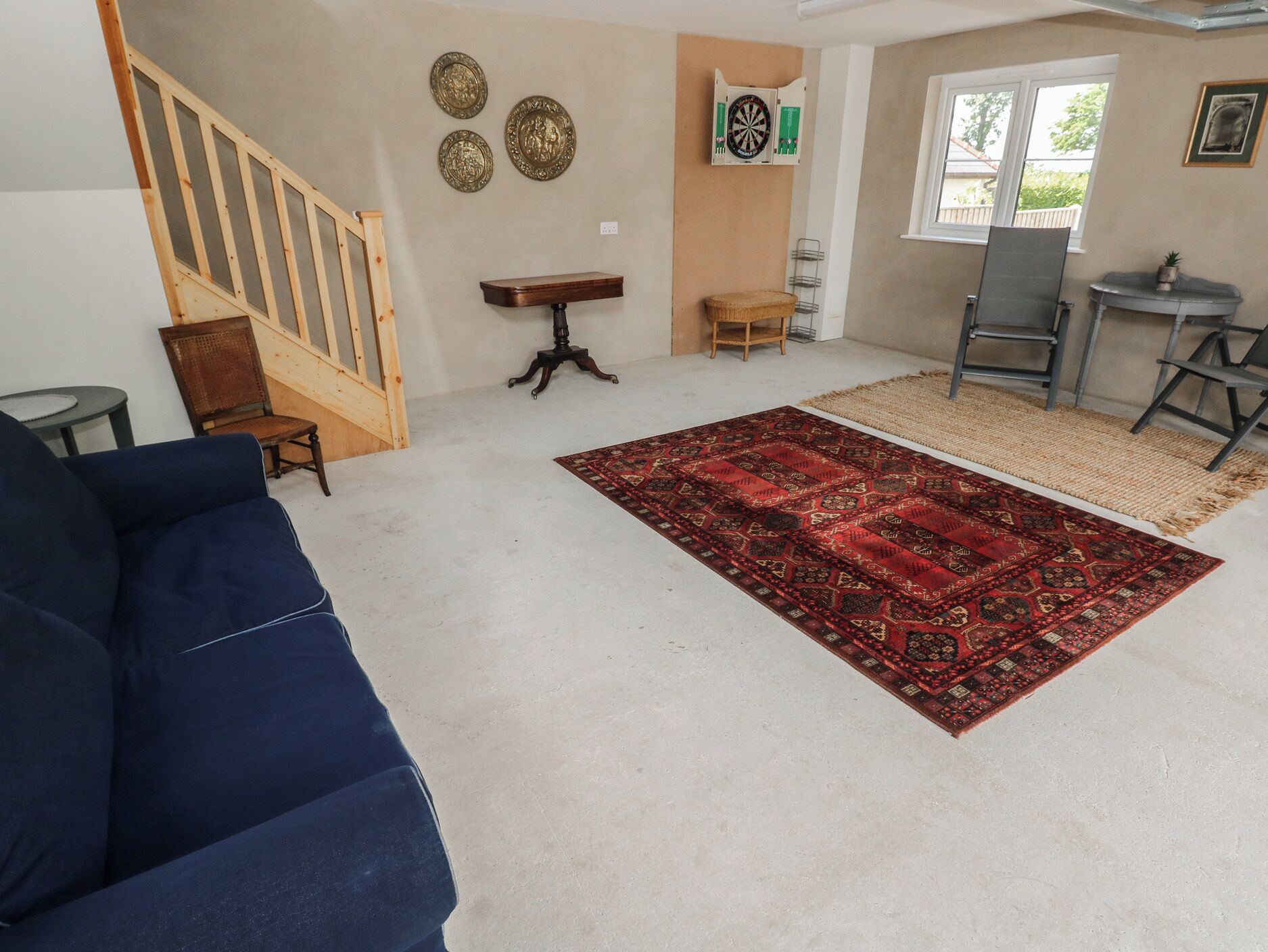 A living room with a blue sofa, a red patterned rug, a wooden staircase, a small table, and a dartboard on the wall.
