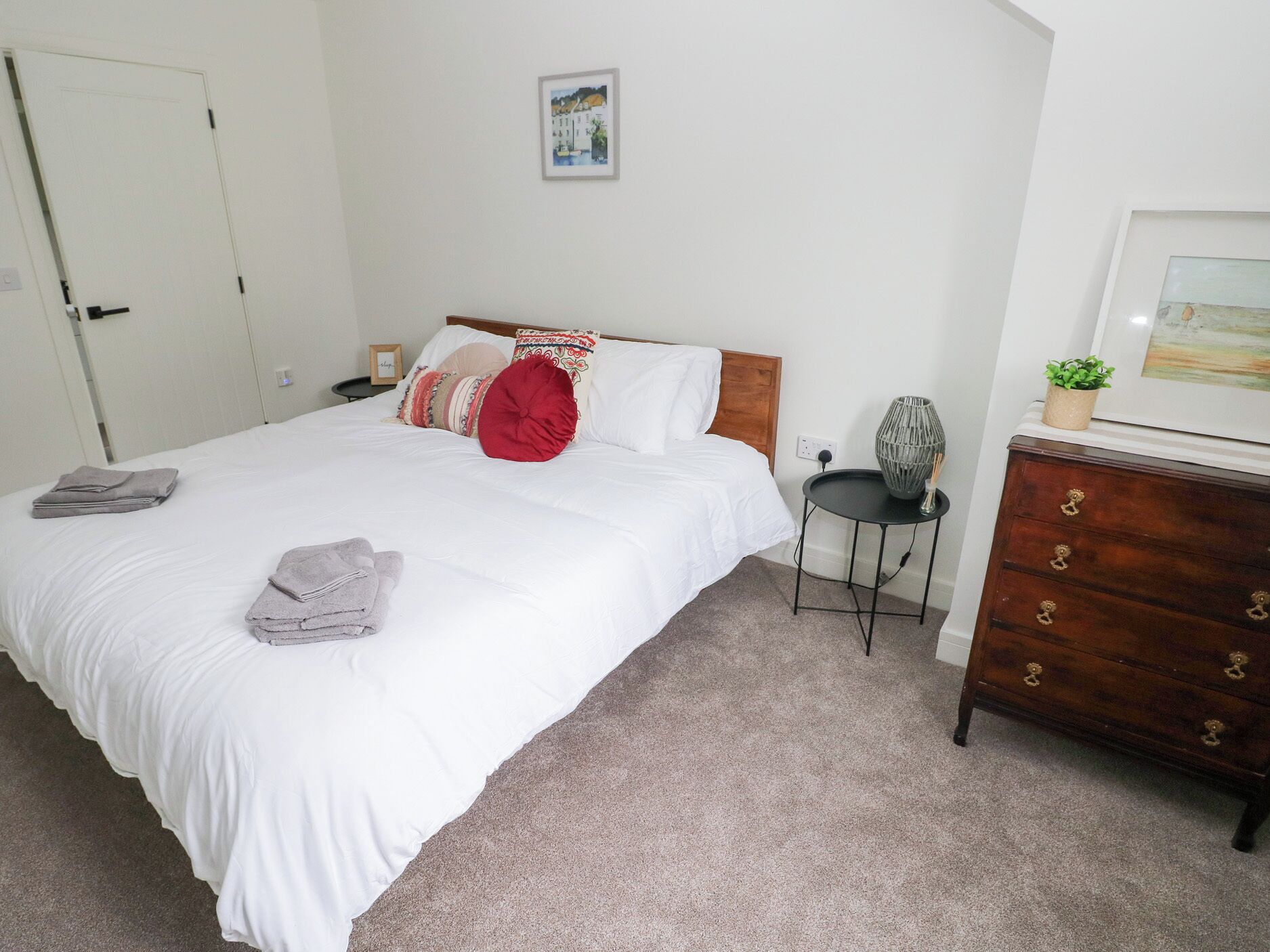 Een net opgemaakt bed met witte lakens en meerdere kussens, een klein nachtkastje met een lamp, een dressoir met lades, en een ingelijst foto aan de muur.