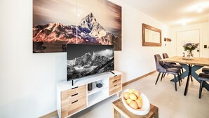 Un salon moderne avec un téléviseur à écran plat fixé sur une console en bois, un coin repas avec une table et des chaises en bois, et un grand tableau représentant un paysage de montagne accroché au mur.