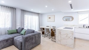 Ein modernes Wohnzimmer mit einer grauen Eckcouch, einem Essbereich mit weißem Tisch und Stühlen sowie einer Küche mit Marmorarbeitsplatte.