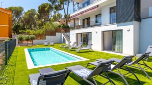 Ein moderner Poolbereich im Freien mit einem rechteckigen Pool, umgeben von Kunstgrün und ausgestattet mit mehreren Liegestühlen und einem Tisch.