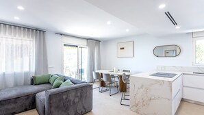 Un salon moderne avec un canapé d’angle, un coin repas avec une table ronde et des chaises, et une cuisine dotée d’un plan de travail en marbre.