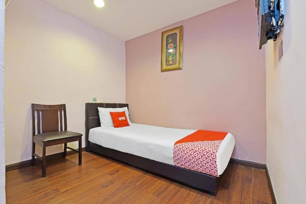 A single bed with a white and red patterned bedspread, a wooden chair, and a framed picture on the wall.