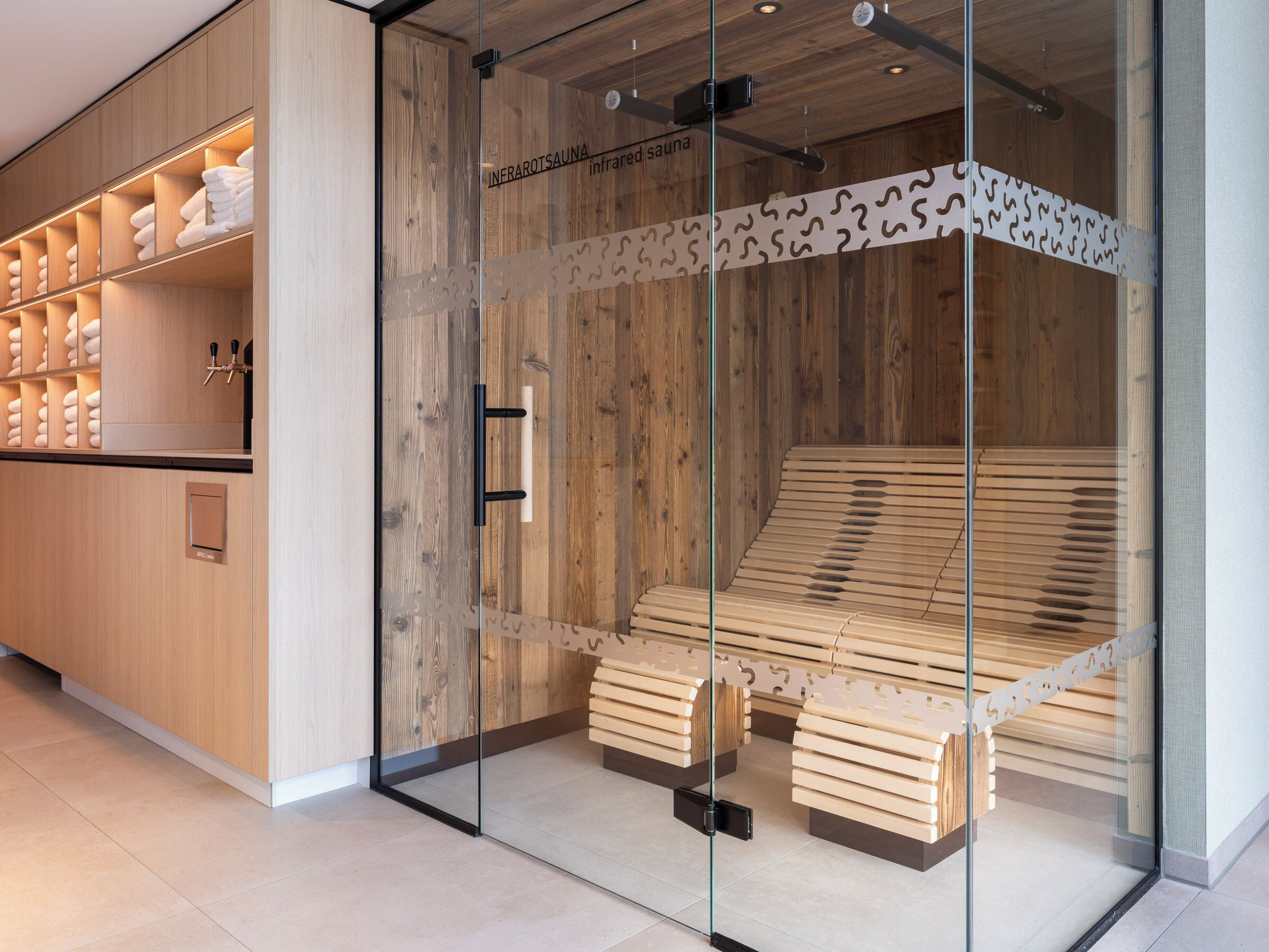 Eine Sauna mit Holzbänken und einer Glastrennwand, einem Handtuchhalter sowie einer Holzwand mit Regalen.
