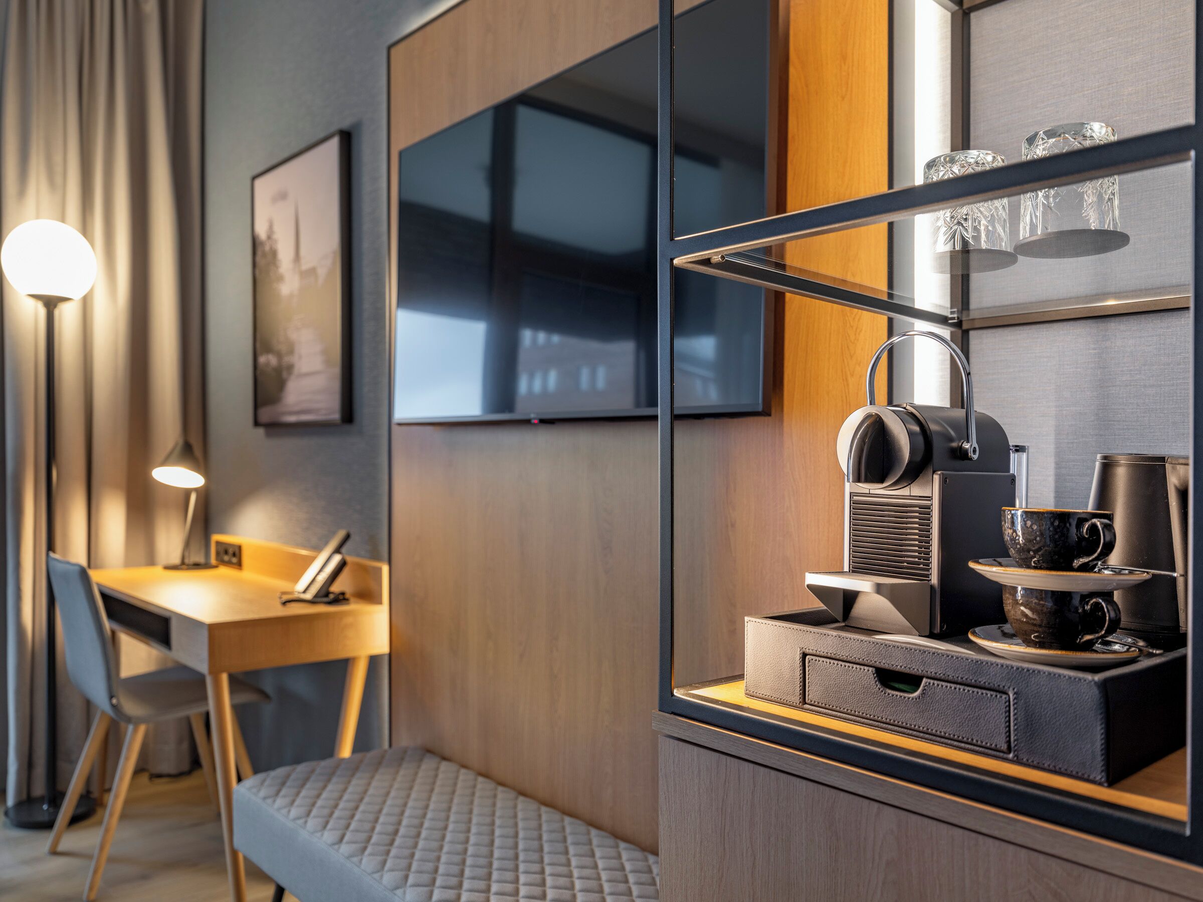 A modern hotel room with a wooden desk, a comfortable chair, a sofa, a coffee machine, and a television mounted on the wall.