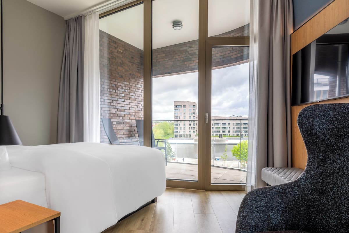 A hotel room with a bed, a chair, a table, and a large window offering a view of buildings and a river.