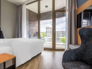 A hotel room with a bed, a chair, a table, and a large window offering a view of buildings and a river. - Scanhotels Stadthafen - Neueröffnung (Rostock)