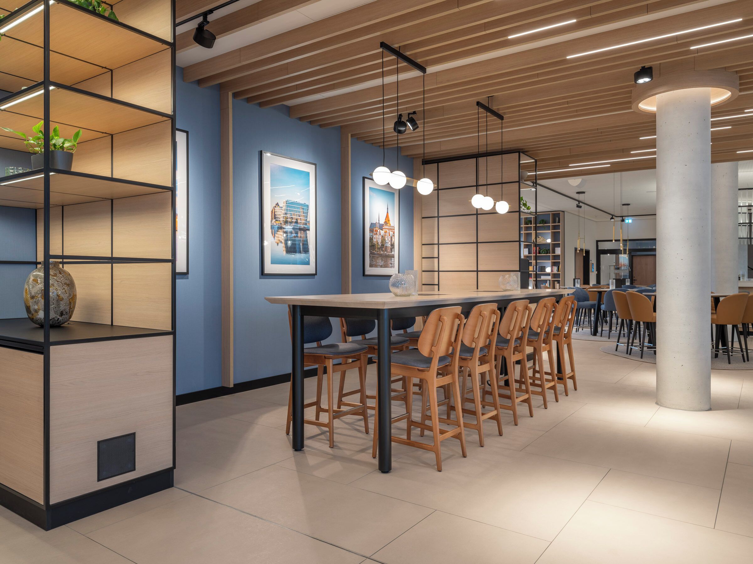 A modern dining area with a long table, chairs, pendant lights, and a blue wall with framed pictures.
