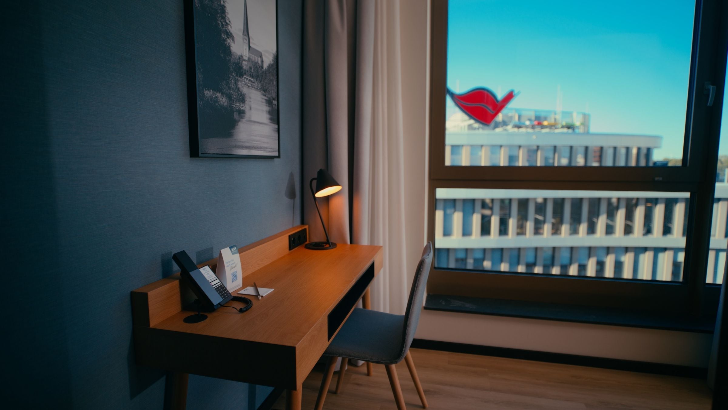 Quarto de hotel com escrivaninha de madeira, cadeira, luminária de mesa, quadro pendurado na parede e janela com vista para edifícios.