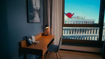 Quarto de hotel com escrivaninha de madeira, cadeira, luminária de mesa, quadro pendurado na parede e janela com vista para edifícios.
