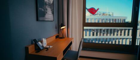 Et hotelværelse med et træskrivebord, en stol, en skrivebordslampe, et billede i ramme på væggen og et vindue med udsigt til bygninger.