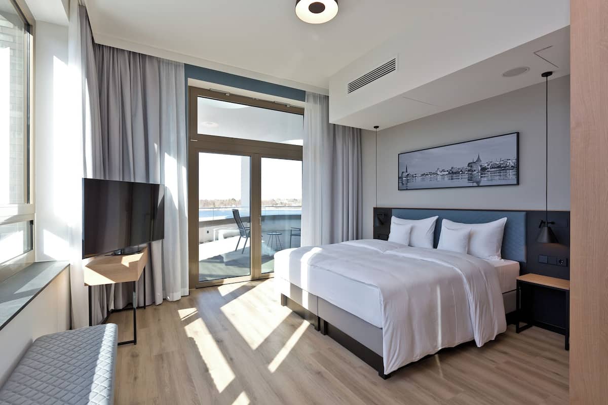 A modern hotel room with a large bed, a TV, a desk, and a balcony with a cityscape view.