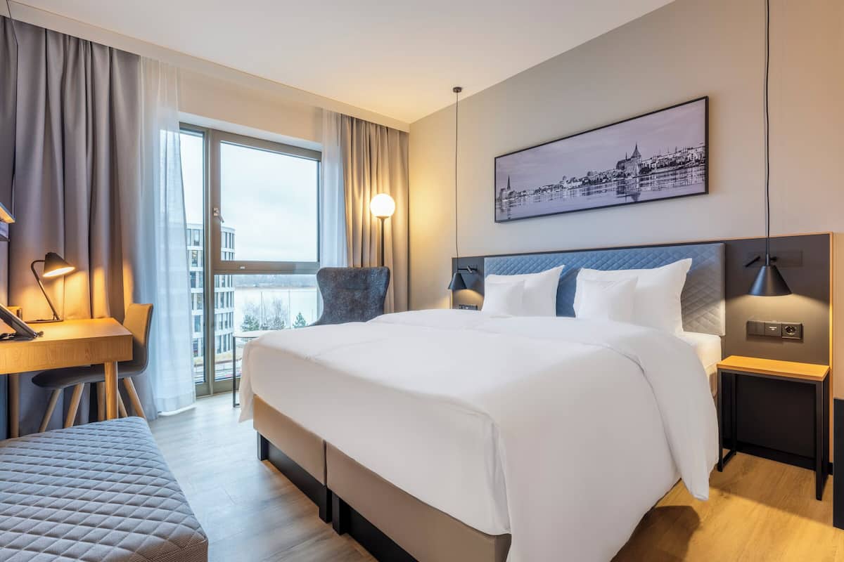 A hotel room with a large bed, a desk, a chair, a bedside table with a lamp, and a cityscape painting on the wall.