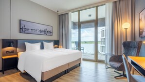 A hotel room with a large bed, a desk with a chair, a view of a body of water, and a cityscape painting on the wall. - Scanhotels Stadthafen - Neueröffnung (Rostock)