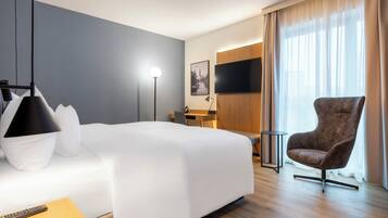 Ein Hotelzimmer mit einem großen Bett, einem Schreibtisch, einem Sessel, einem Fernseher und einem Fenster mit Vorhängen.