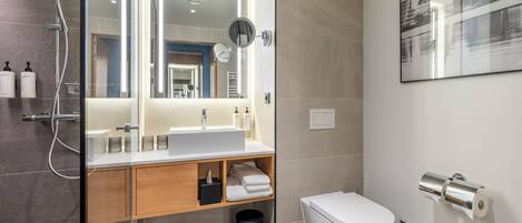 A modern bathroom with a glass-enclosed shower, a wooden vanity, a white sink, a mirror, a toilet, and a roll of toilet paper.