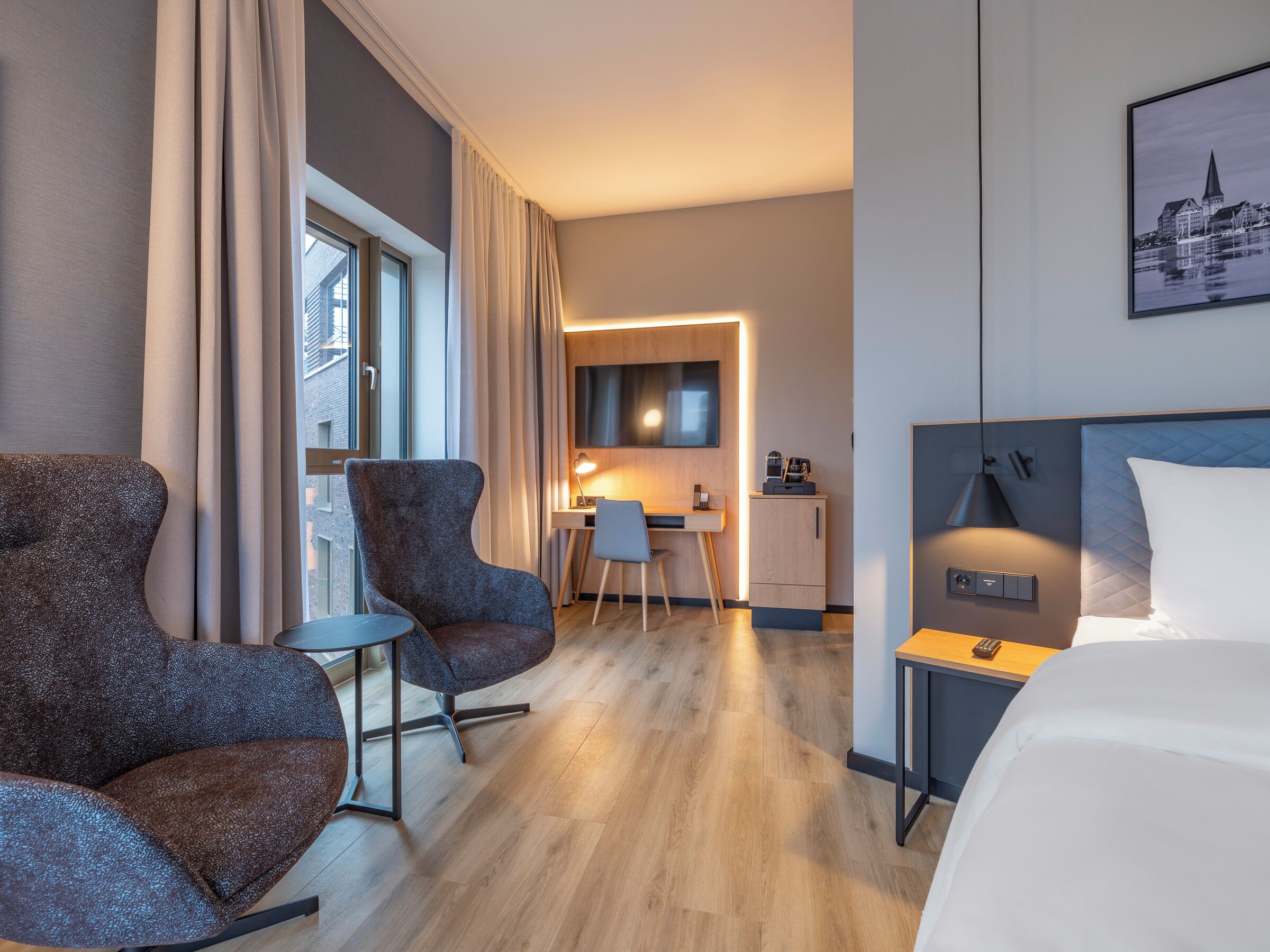 A hotel room with a bed, two armchairs, a desk with a chair, a small cabinet, and a window with curtains.