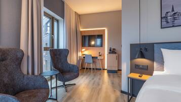 Ein Hotelzimmer mit einem Bett, zwei Sesseln, einem Schreibtisch mit Stuhl, einer kleinen Kommode und einem Fenster mit Vorhängen.
