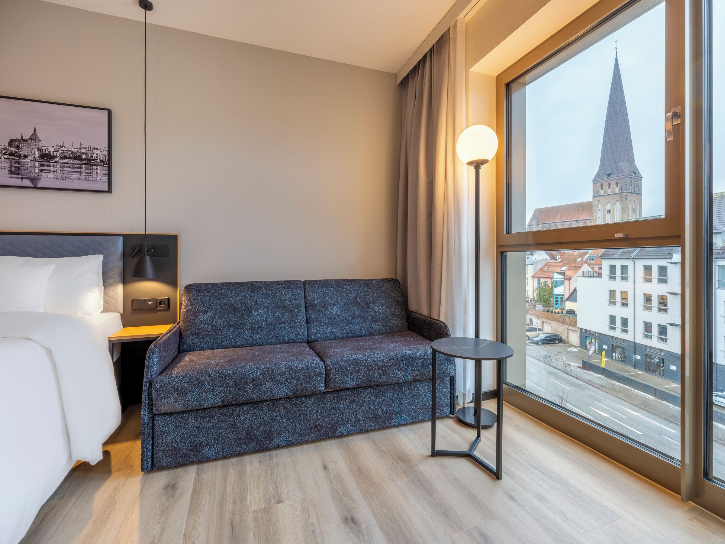 A modern hotel room with a bed, a grey sofa, a small table, and a large window offering a city view.